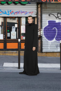 Person wearing a long black dress standing on a city sidewalk in front of graffiti-covered shop shutters and posters about the storefronts.