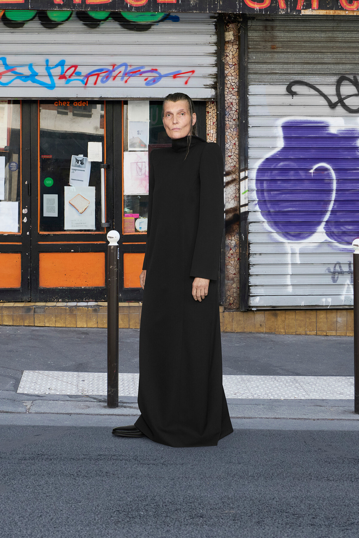Person wearing a long black dress standing on a city sidewalk in front of graffiti-covered shop shutters and posters about the storefronts.
