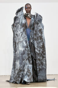 Model on runway wearing a dramatic metallic, crinkled-gray sculptural gown with an off-center deep V, blue accents, and high collar accessory, neutral background.