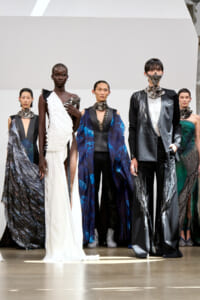 Group of diverse models on a runway wearing avant-garde gowns and metallic fabrics, posing for a fashion show.