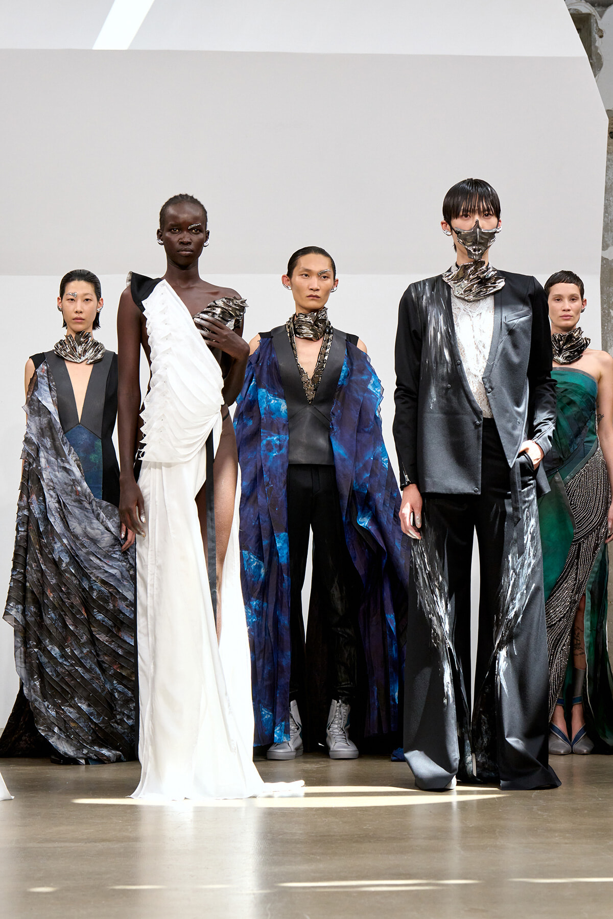 Group of diverse models on a runway wearing avant-garde gowns and metallic fabrics, posing for a fashion show.