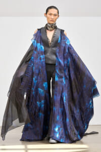 Model on a runway wearing a dark metallic vest under a voluminous blue-black sheer cape with pleated panels.