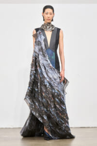 Female model on a runway wearing a sleeveless black gown with a deep V, metallic collar, and a dramatic, layered marbled scarf draped over one shoulder and down the length of the dress.