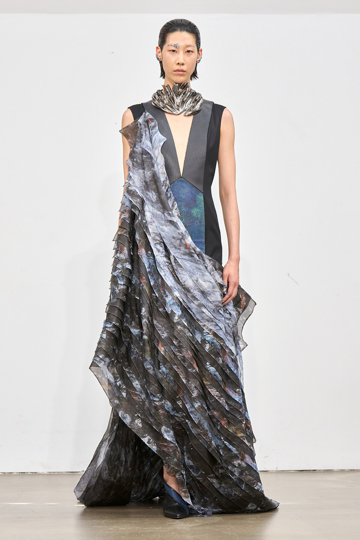 Female model on a runway wearing a sleeveless black gown with a deep V, metallic collar, and a dramatic, layered marbled scarf draped over one shoulder and down the length of the dress.