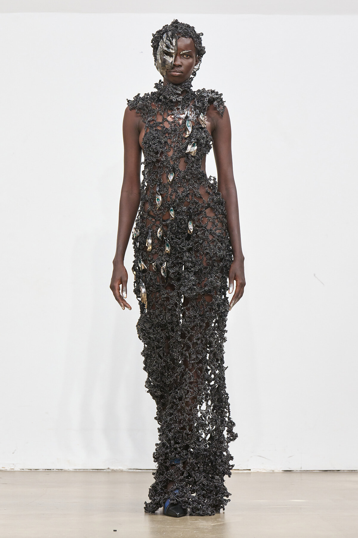 Model on a runway wearing a full-length, dark, open-knit gown with metallic shell embellishments and a matching headpiece.