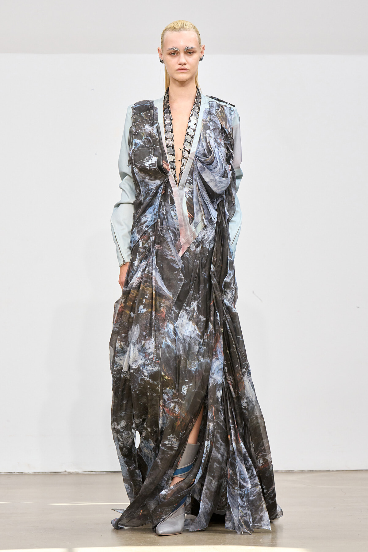 Runway model in a long metallic gown with pale blue sleeves, featuring a deep V-neck and draped, marbled fabric.