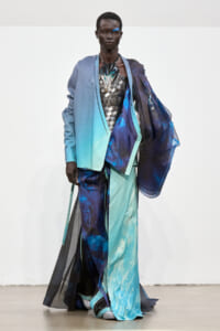 Runway model in a gradient blue-teal avant-garde coat with sheer panels over a metallic bodice and flowing layers.