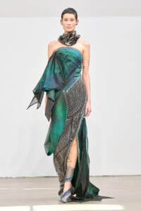 Runway model wearing a strapless green and black draped gown with a metallic sculpted neckpiece and flowing beaded skirt.