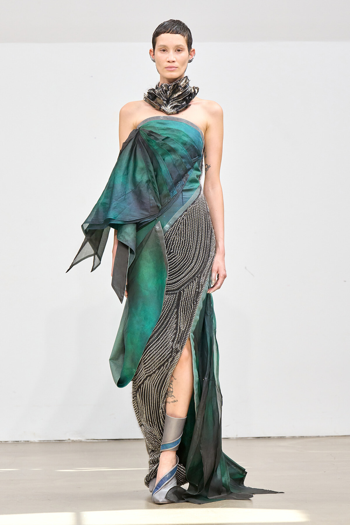 Runway model wearing a strapless green and black draped gown with a metallic sculpted neckpiece and flowing beaded skirt.
