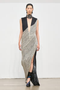 Runway model in a silver chainmail gown with black panel shoulders and a deep V-neck, finished with a front slit and ankle boots.
