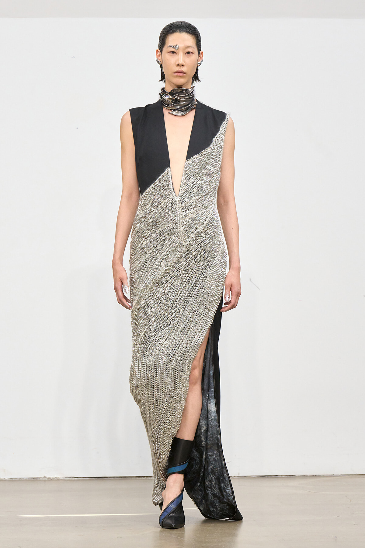 Runway model in a silver chainmail gown with black panel shoulders and a deep V-neck, finished with a front slit and ankle boots.