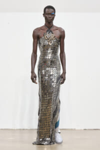 Male model on a runway wearing a metallic lattice gown with a gold leaf chest embellishment and beaded fringe, paired with white boots.