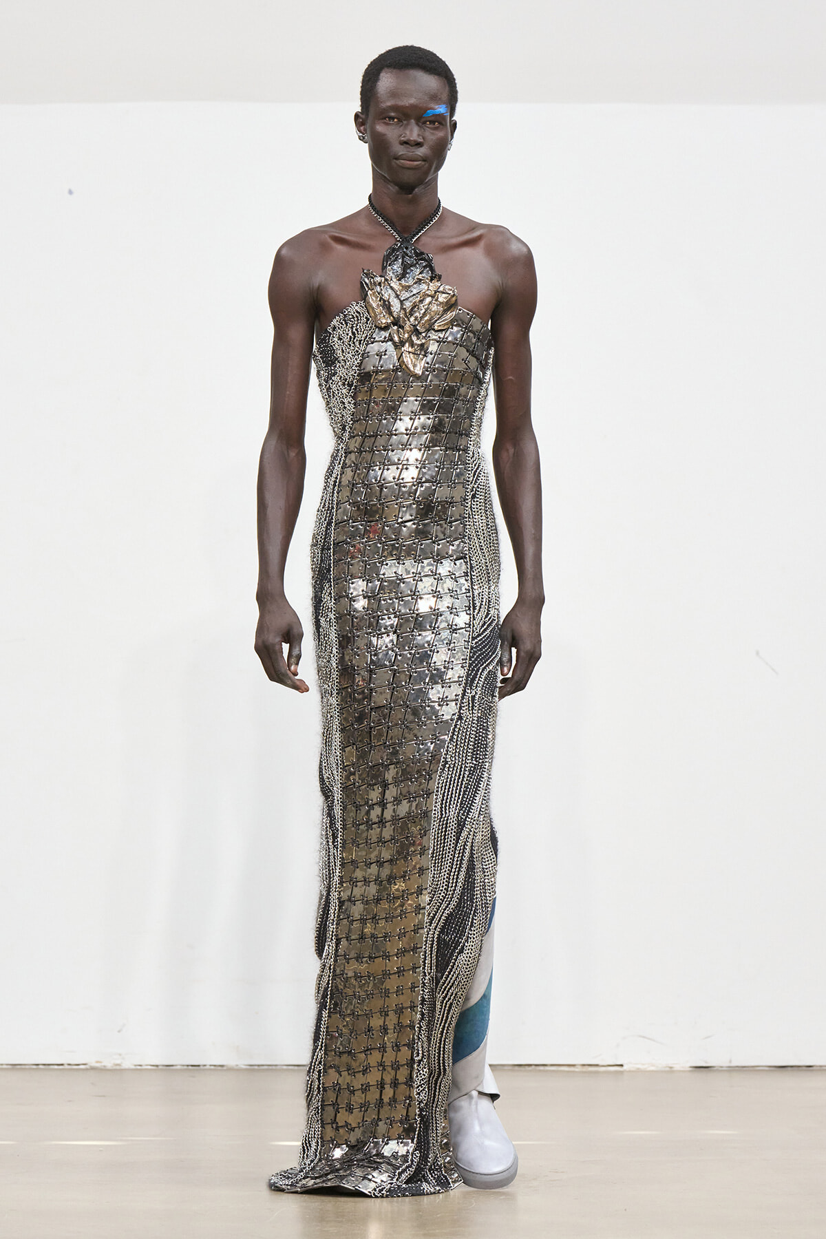 Male model on a runway wearing a metallic lattice gown with a gold leaf chest embellishment and beaded fringe, paired with white boots.