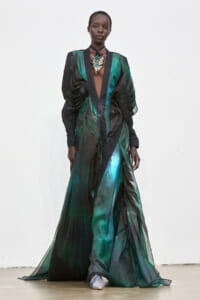 Model wearing a floor-length green and black sheer gown with dramatic ruched sleeves and a metallic leaf pendant on a white runway background, front view.