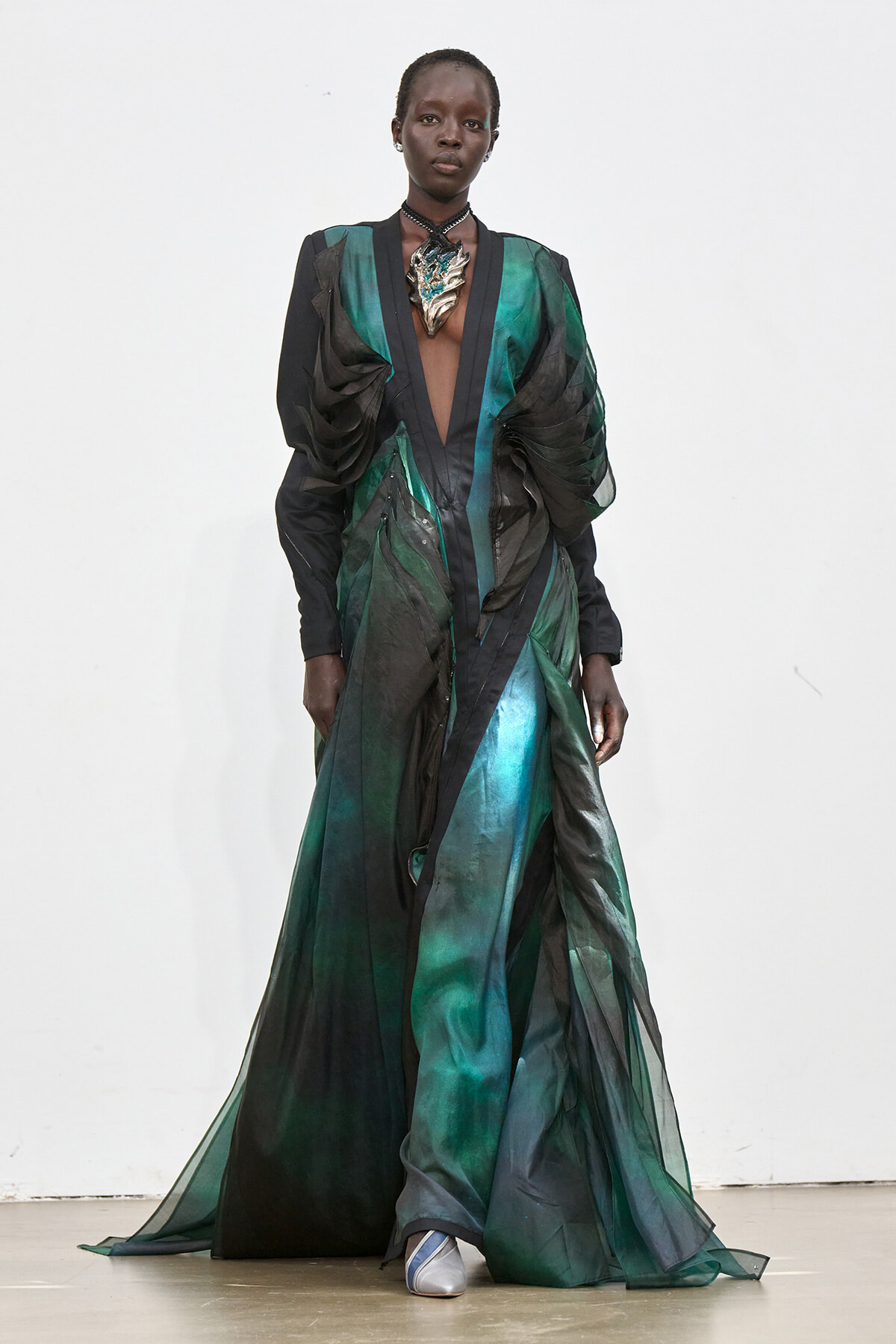 Model wearing a floor-length green and black sheer gown with dramatic ruched sleeves and a metallic leaf pendant on a white runway background, front view.