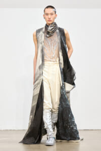 Male runway model wearing an avant-garde metallic sleeveless coat over a sheer lace top and high-waisted beige trousers, with a sculptural scarf and gray boots.