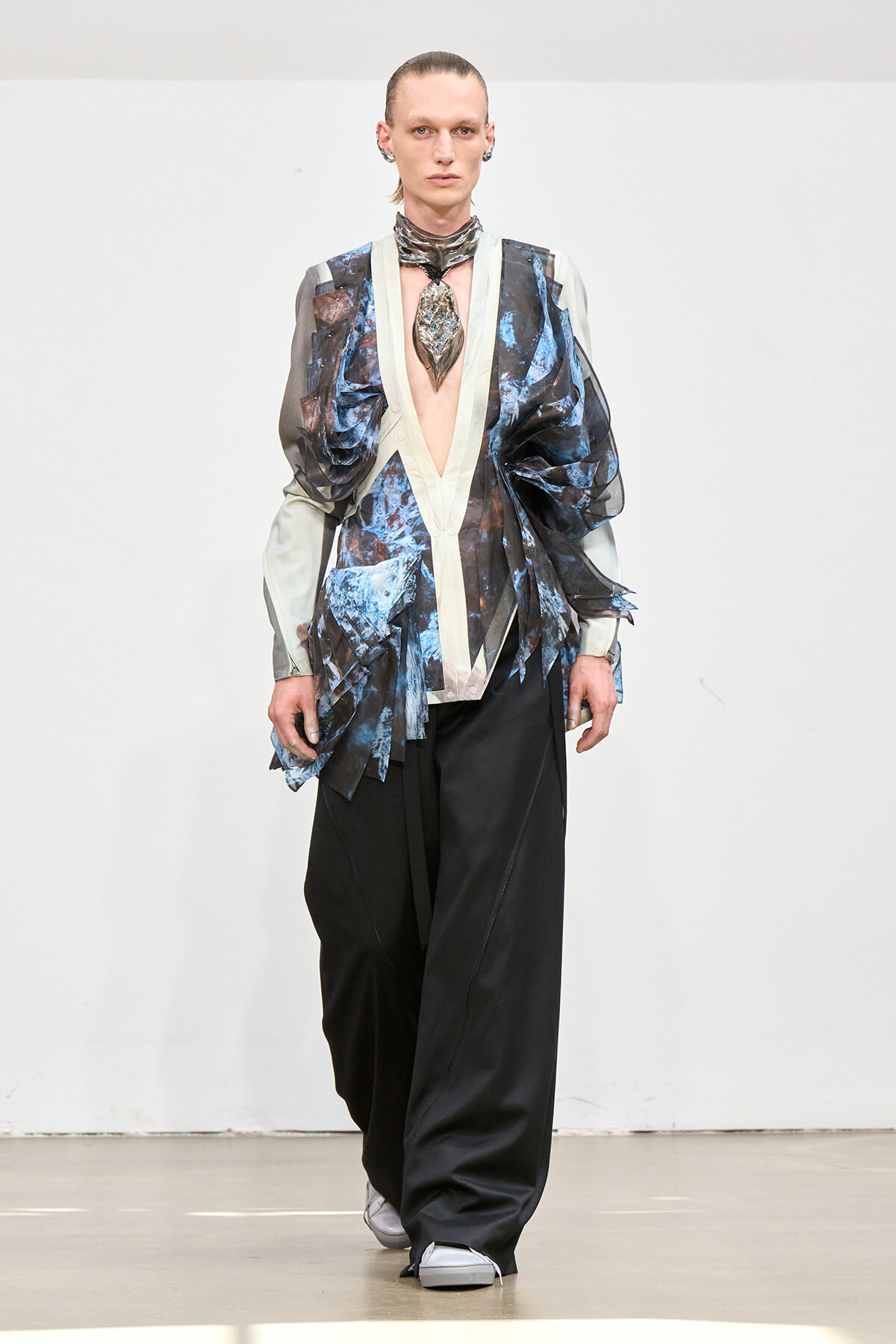 Model on a runway wearing a deep-plunge sheer-sleeve jacket with blue-black abstract print, beige trim, and wide black pants.