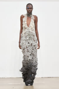 Model walks the runway in a sleeveless, floor-length lace gown that fades from cream at the top to black at the bottom with a textured, openwork pattern.