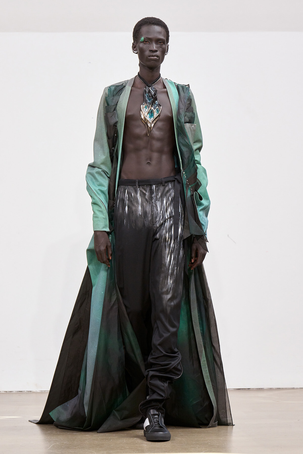 Male model on a runway wearing a long green and black sheer cape, open chest revealing a metallic pendant necklace and glossy black pants with high boots.