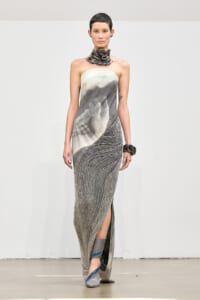Model on runway wearing a strapless gown with a grayscale hands print, chain necklace, and metallic beaded skirt.