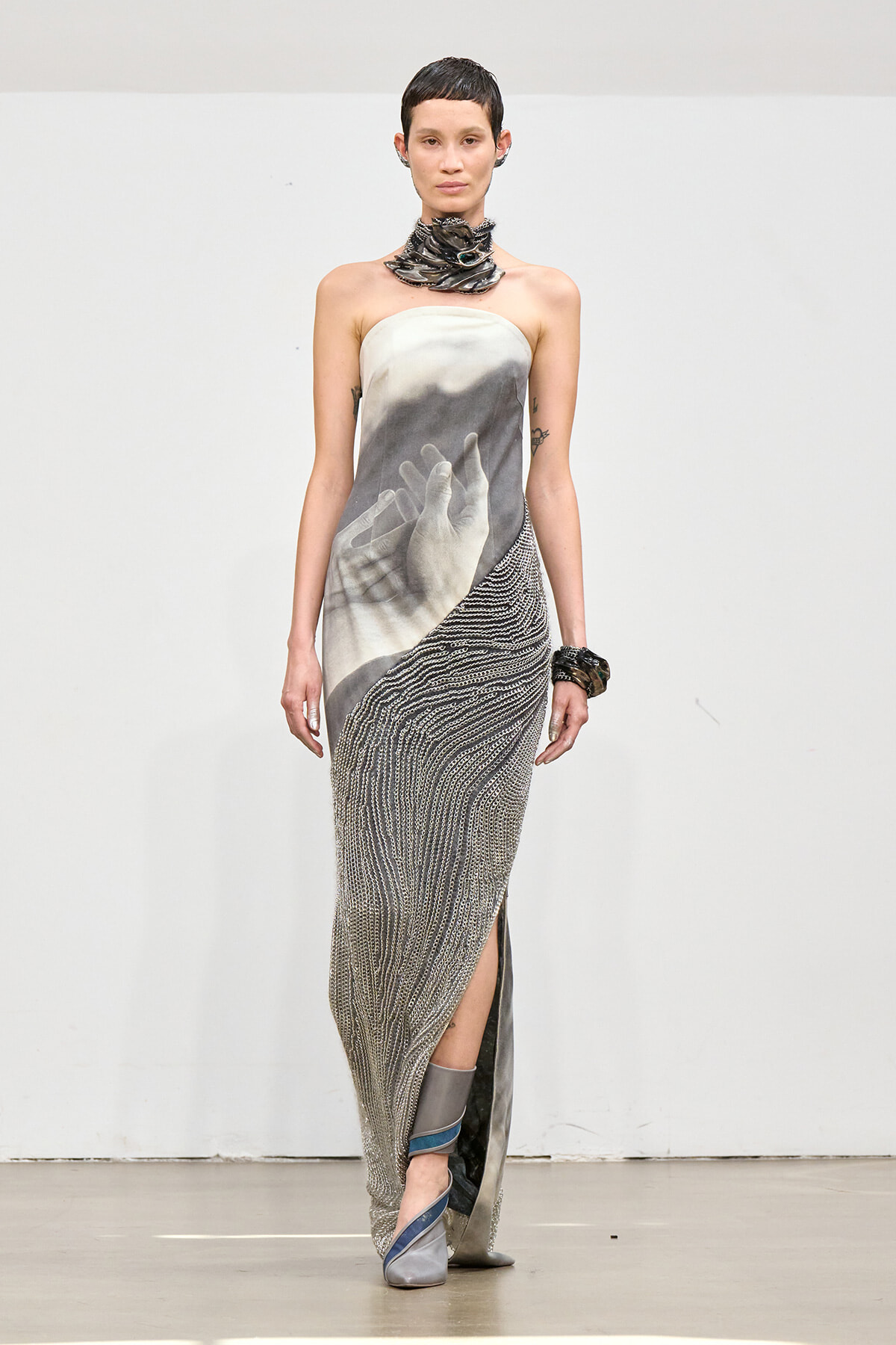 Model on runway wearing a strapless gown with a grayscale hands print, chain necklace, and metallic beaded skirt.