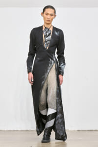 Model on a runway wearing a black, long-sleeved gown with a deep V neckline and a high slit, paired with metallic-patterned tights and boots.