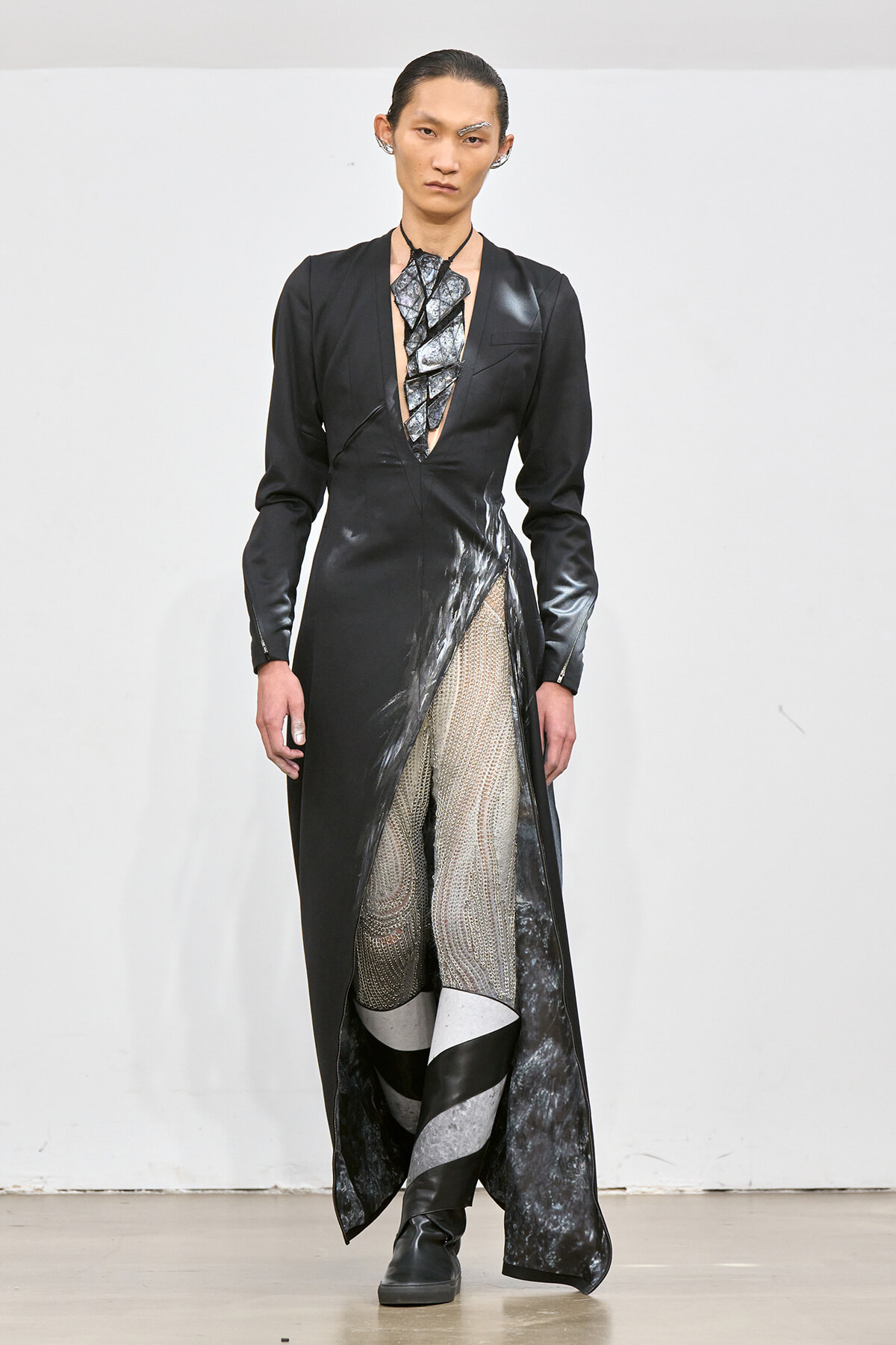 Model on a runway wearing a black, long-sleeved gown with a deep V neckline and a high slit, paired with metallic-patterned tights and boots.