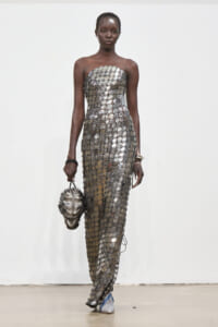 Model on a runway wearing a strapless metallic grid dress, carrying a metallic sculpted handbag resembling a head.