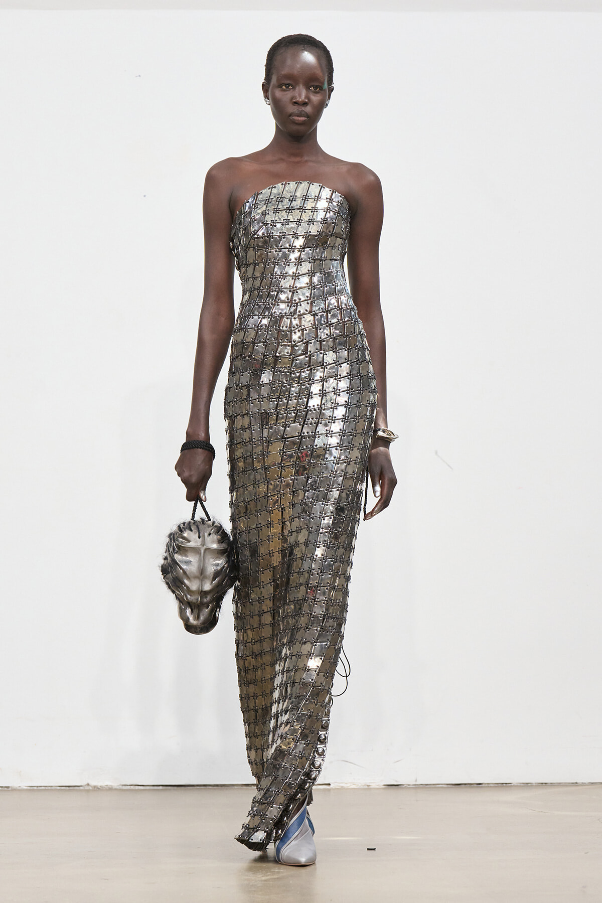 Model on a runway wearing a strapless metallic grid dress, carrying a metallic sculpted handbag resembling a head.