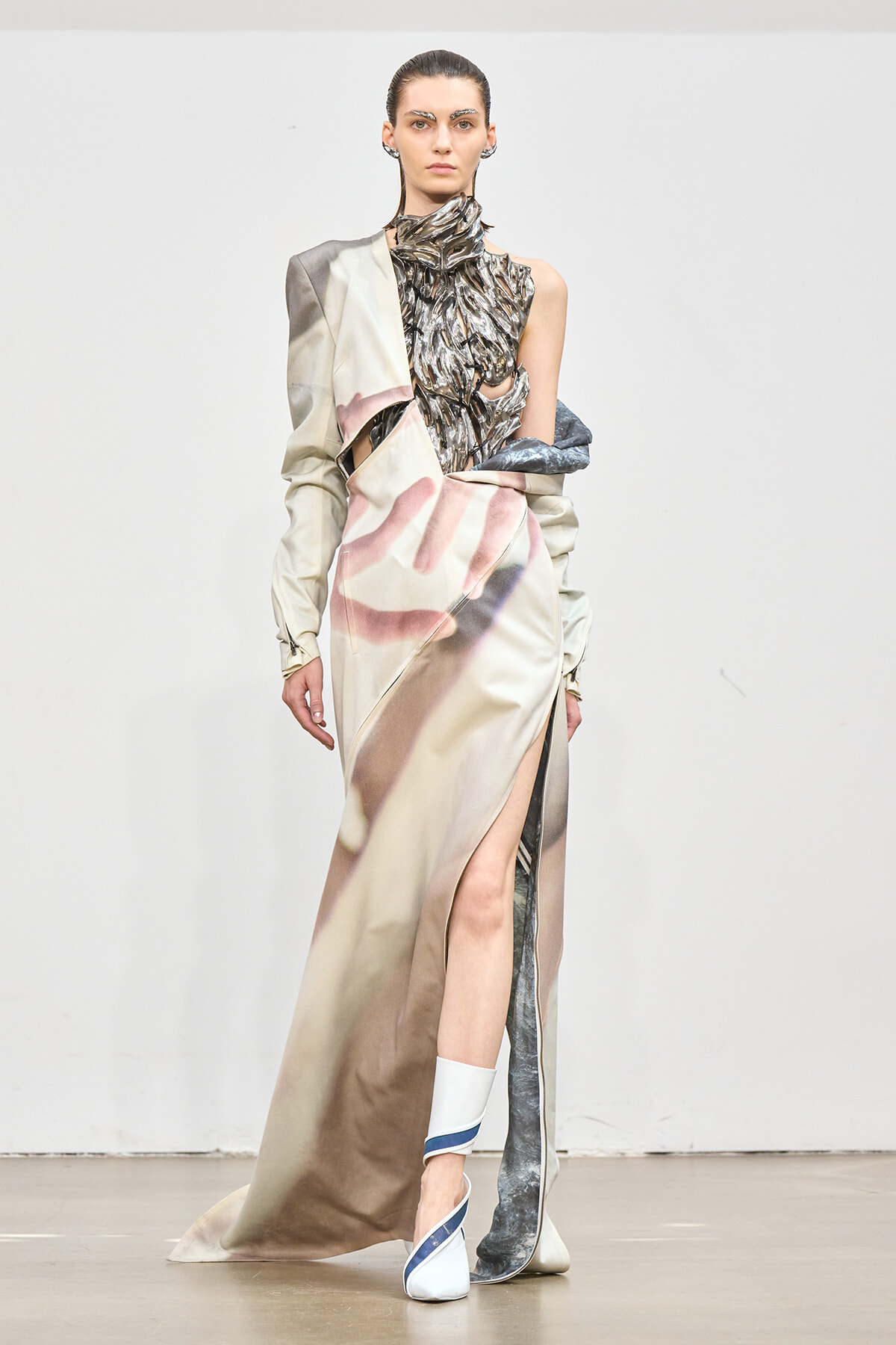 Runway model in a two-tone beige gown with a metallic, ruched bodice, one bare shoulder, and a high thigh slit, paired with white-blue heeled sandals.