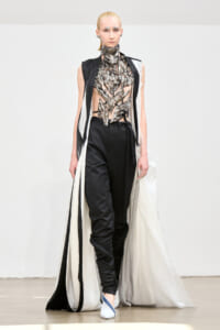 Model on a runway wearing a metallic leaf-inspired bodice, black pants, and a long white cape with black trim.