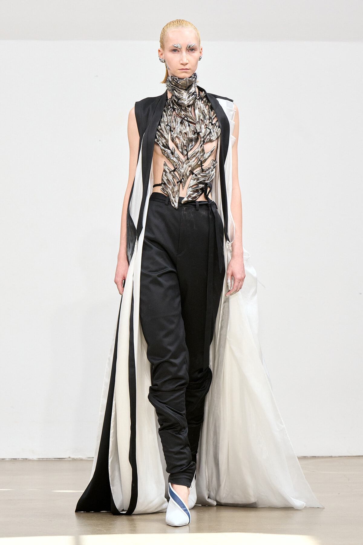 Model on a runway wearing a metallic leaf-inspired bodice, black pants, and a long white cape with black trim.