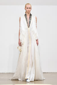 Model on a runway wearing a flowing ivory-white gown with a deep V neckline and black patterned scarf detail at the neck.