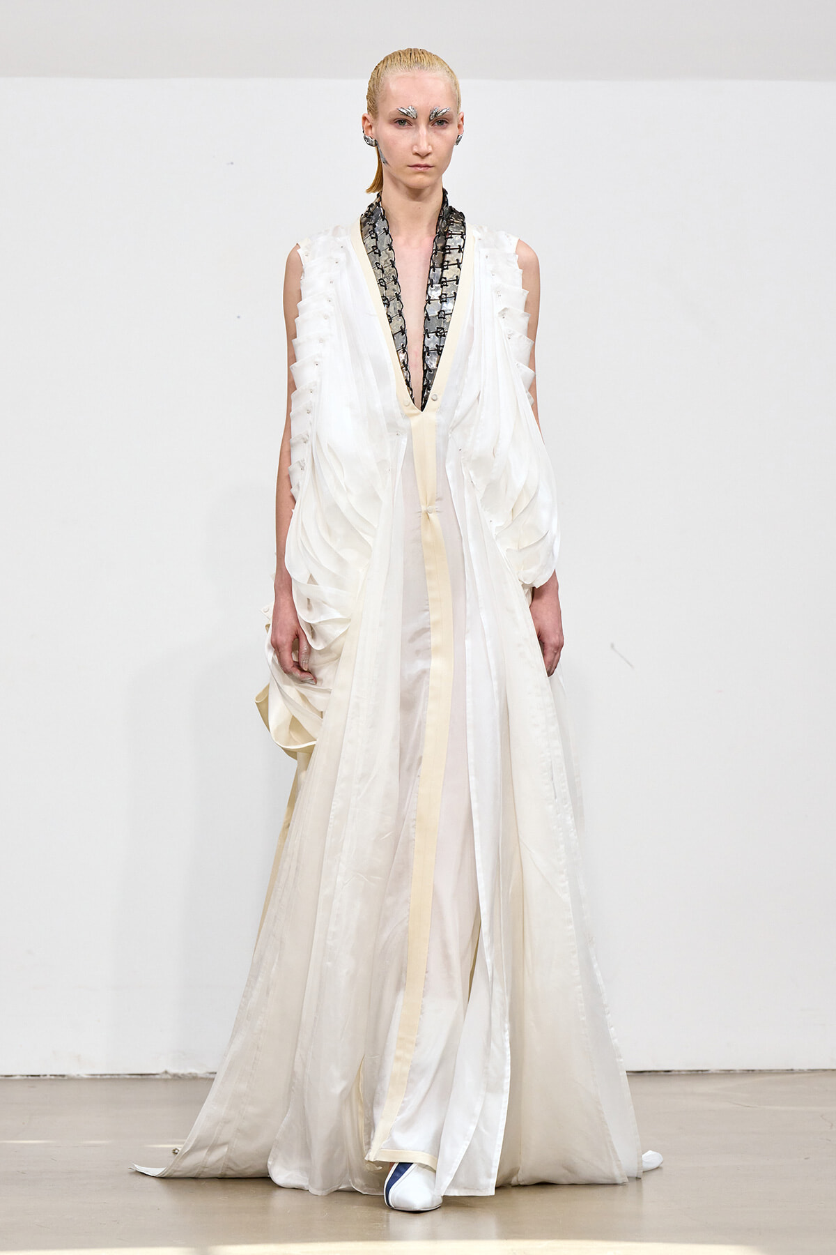 Model on a runway wearing a flowing ivory-white gown with a deep V neckline and black patterned scarf detail at the neck.