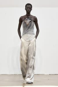 Dark-skinned model poses in a silver beaded strapless top with a sculpted leaf motif, paired with wide, pale silver pants against a neutral backdrop.