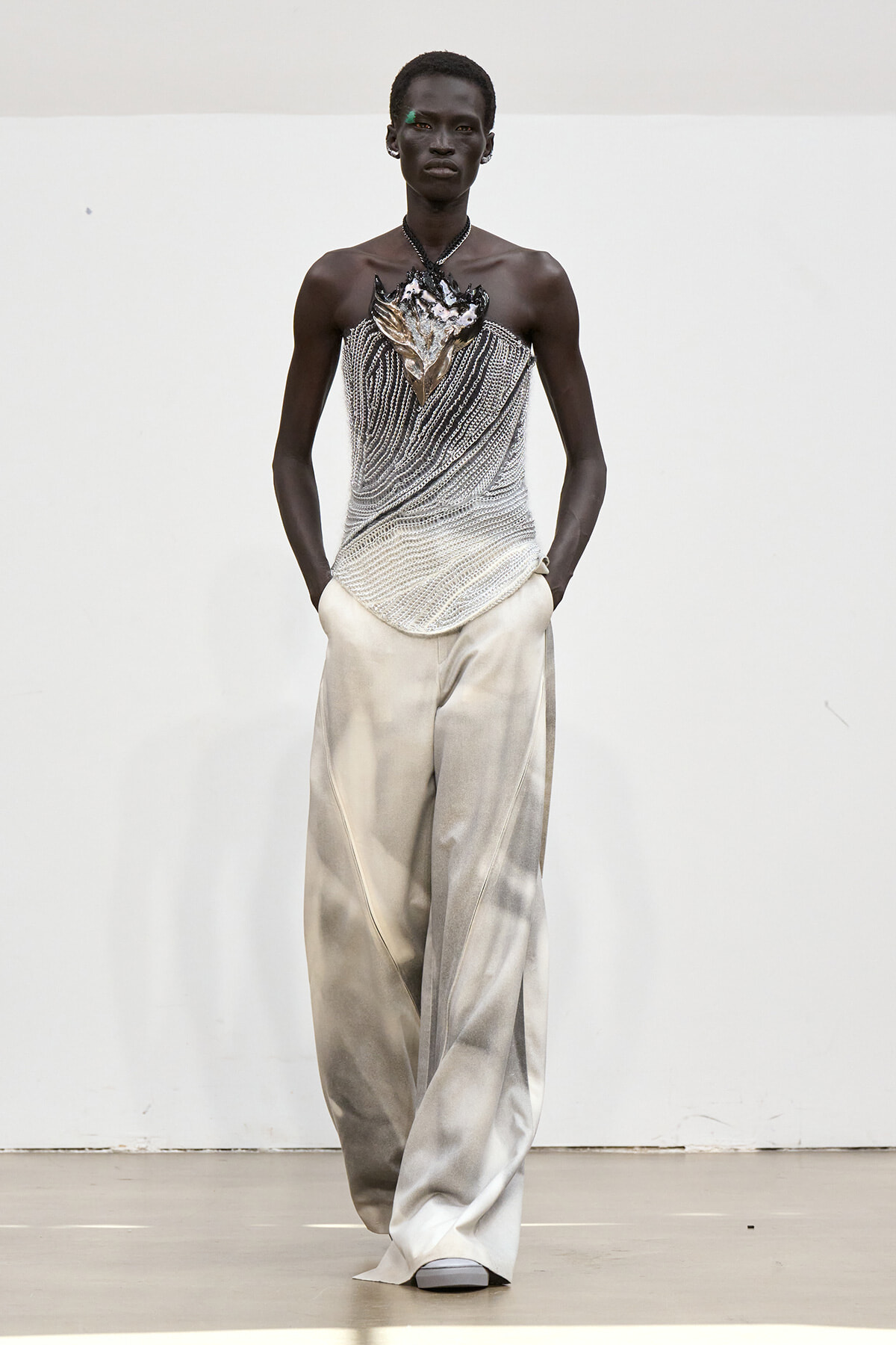 Dark-skinned model poses in a silver beaded strapless top with a sculpted leaf motif, paired with wide, pale silver pants against a neutral backdrop.