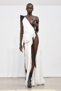 Dark-skinned model walks runway in a white one-shoulder draped gown with black accents, high slit, and blue-black strapped heels.