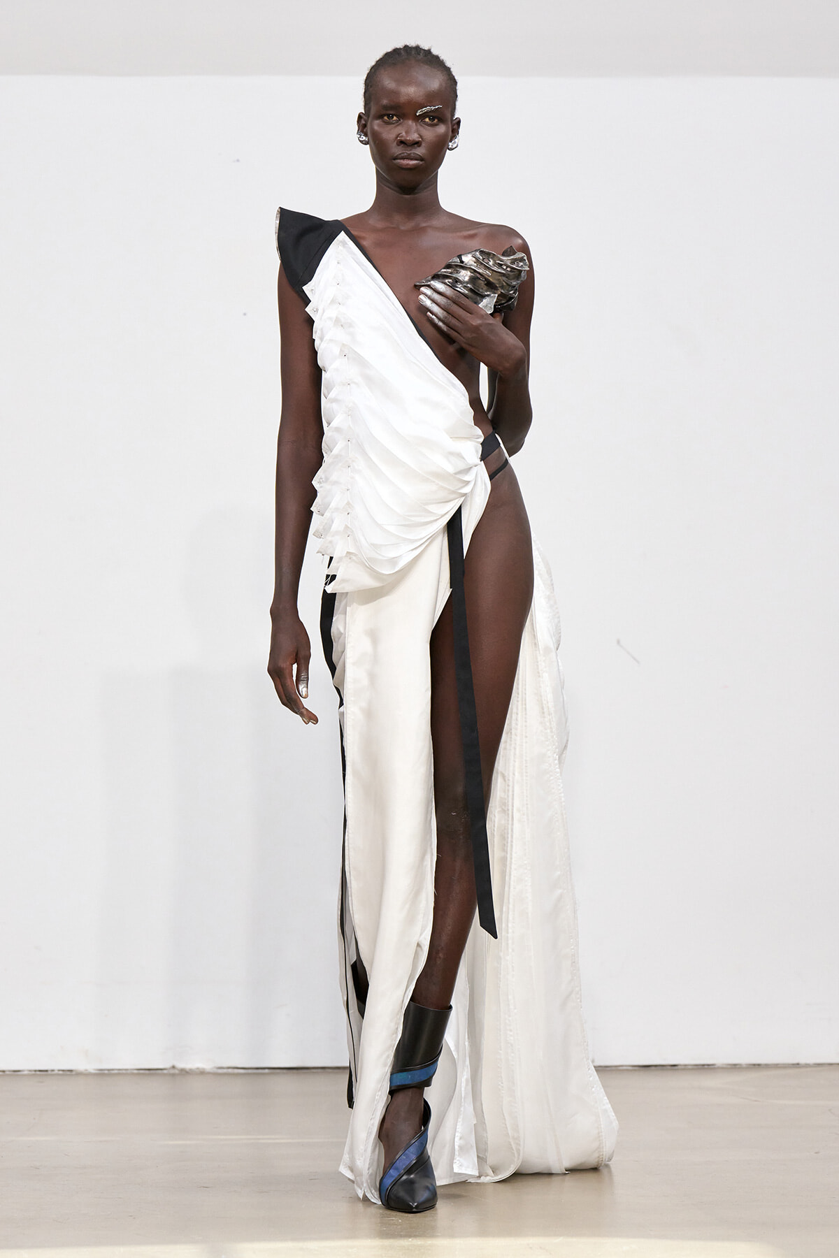 Dark-skinned model walks runway in a white one-shoulder draped gown with black accents, high slit, and blue-black strapped heels.