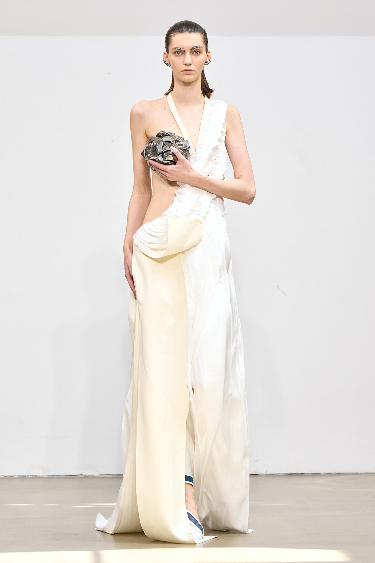 Model on a runway wearing a white, asymmetric draped gown while holding a metallic sculpted clutch near her chest.