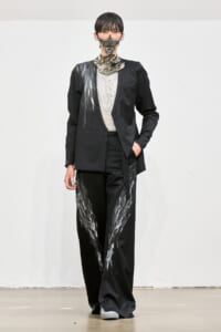 Runway model in a black tailored suit with silver paint streaks on the jacket and wide-leg pants, lace top, and a metallic mask.