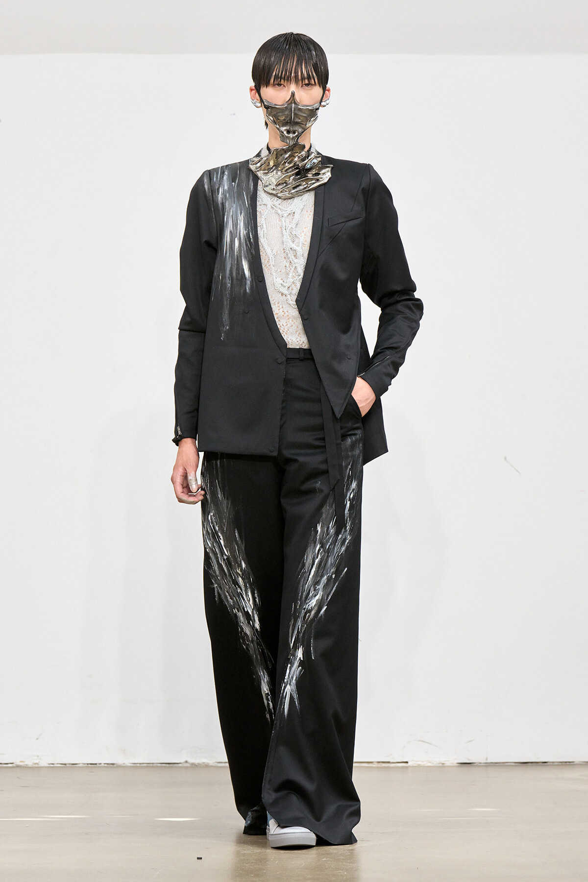 Runway model in a black tailored suit with silver paint streaks on the jacket and wide-leg pants, lace top, and a metallic mask.
