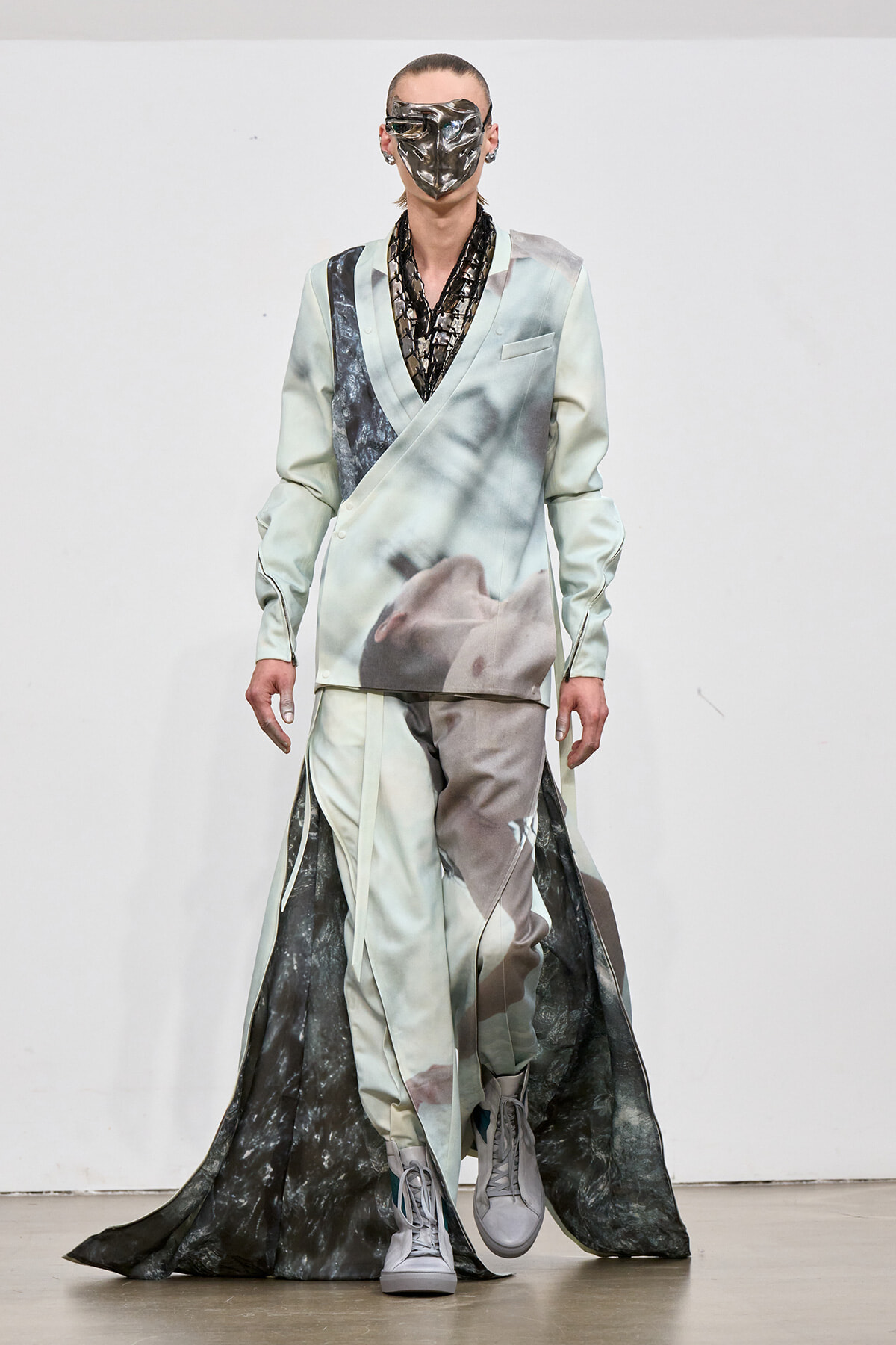 Model on runway in a pale satin double-breasted suit with a large print, matching pants, and a long dark cape; wearing a metallic face mask.