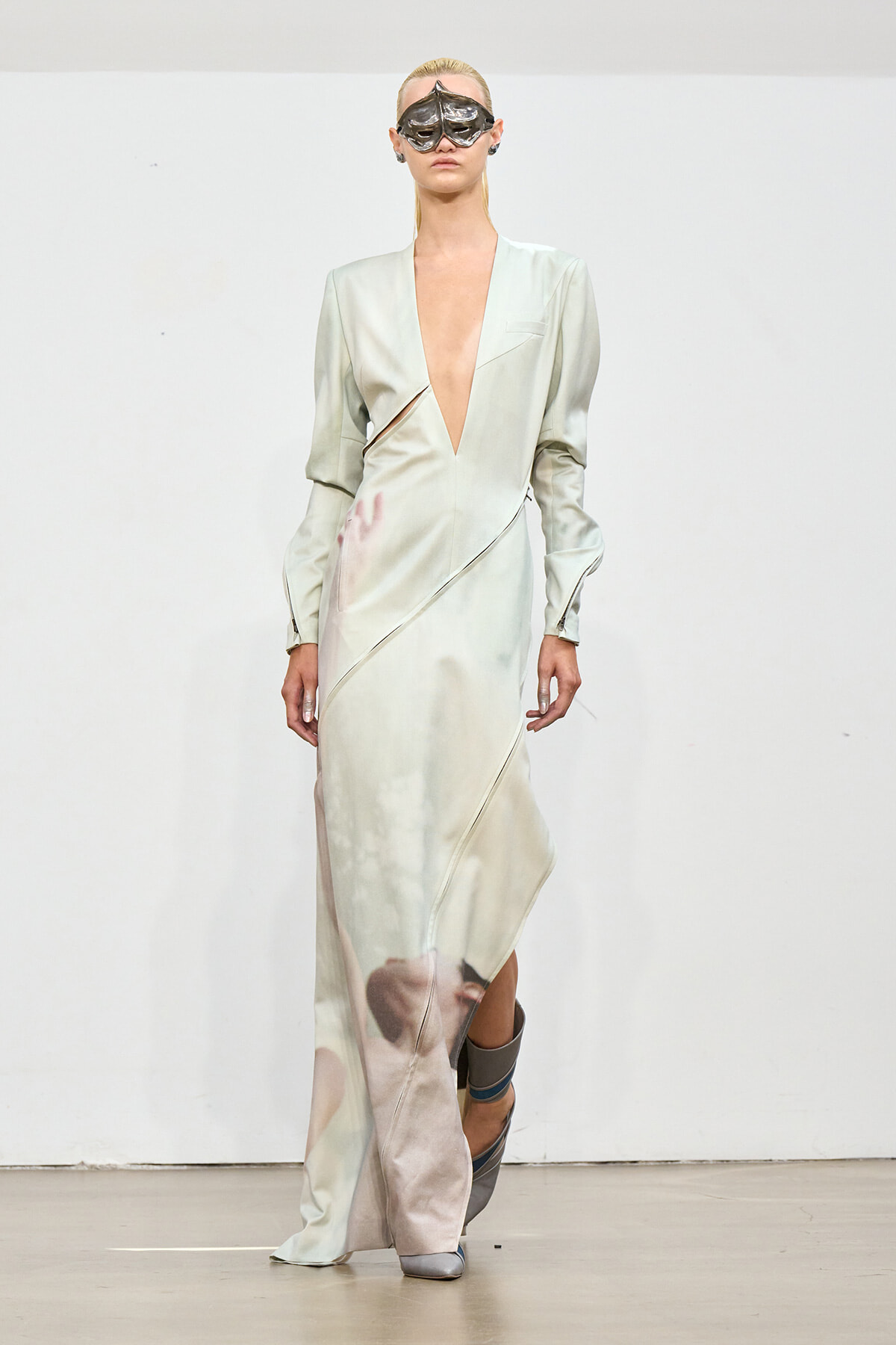 Model on a runway wearing a pale satin gown with a plunging neckline and diagonal seam details, plus a metallic mask.