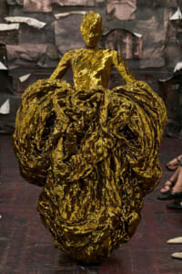 Model walking a runway in a dramatic gold metallic gown with a voluminous, crinkled skirt.