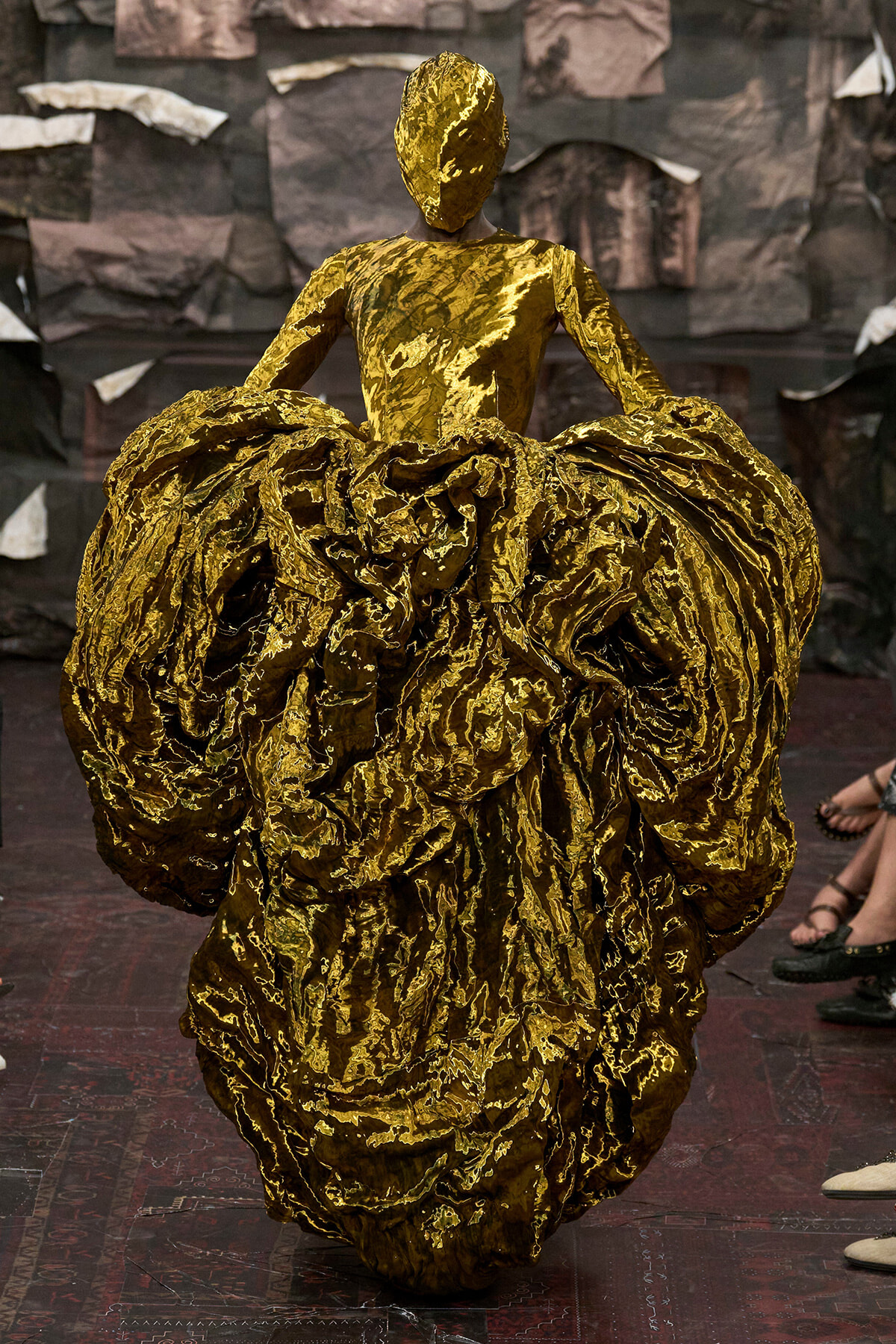Model walking a runway in a dramatic gold metallic gown with a voluminous, crinkled skirt.