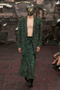 Runway model in a dark green, heavily beaded suit with an open jacket and no shirt, paired with a metallic headpiece/mask.