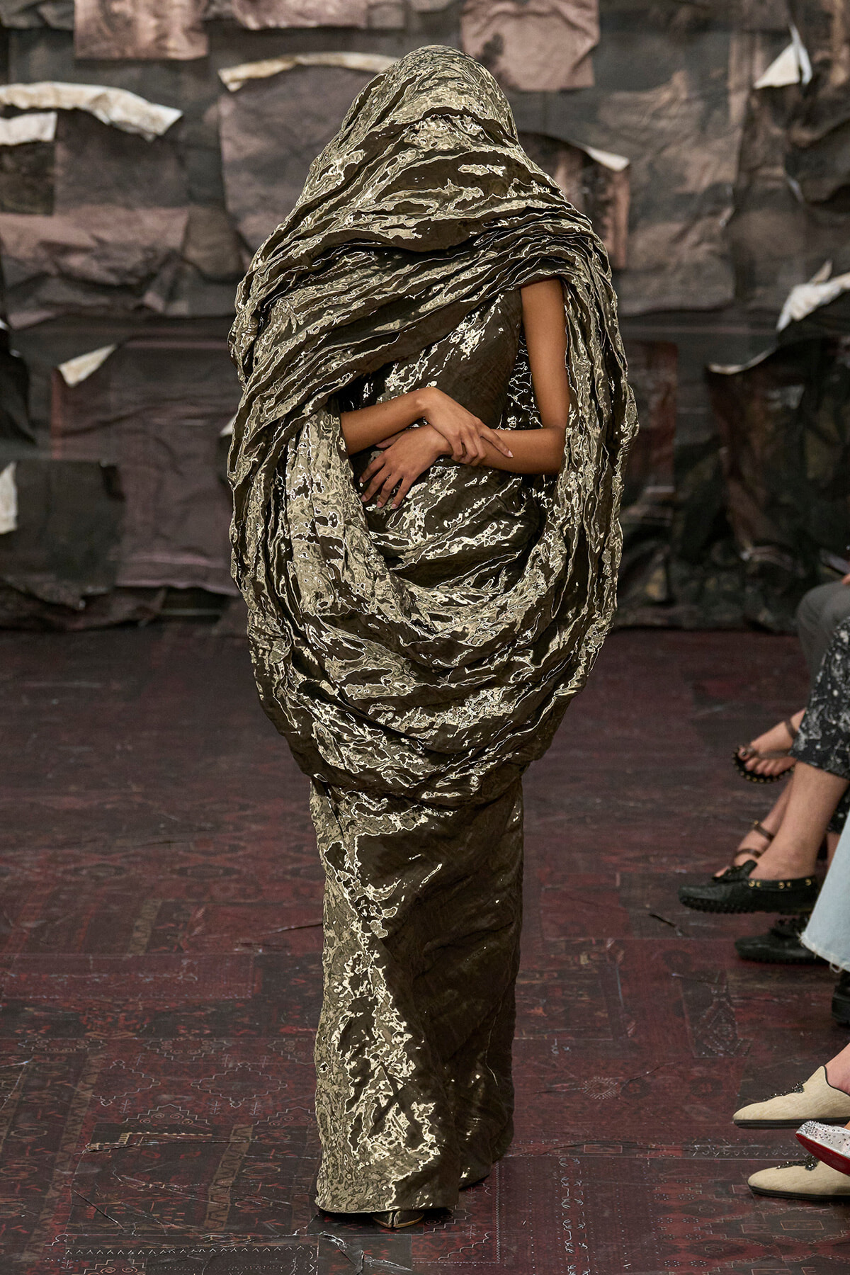 Model walks the runway in a dramatic, oversized wrap dress with a hooded, layered metallic-brown fabric and abstract white marbling.
