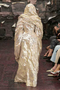 Back view of a model in a crinkled metallic beige hooded gown walking a runway, with seated audience nearby.