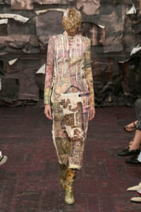 Runway model wearing a full-body patchwork dress crafted from torn fabrics in muted pinks and browns, with a shredded fabric head covering and matching boots.