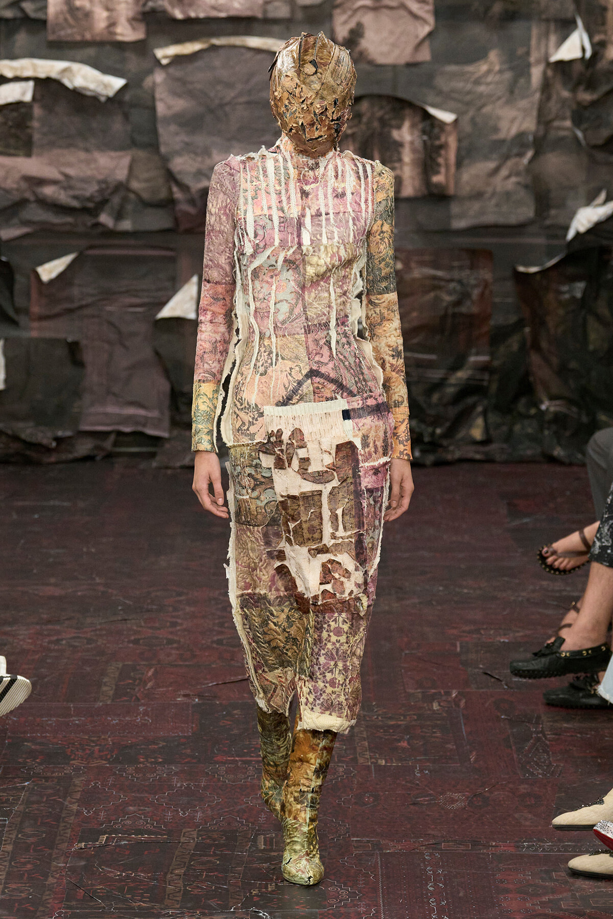Runway model wearing a full-body patchwork dress crafted from torn fabrics in muted pinks and browns, with a shredded fabric head covering and matching boots.